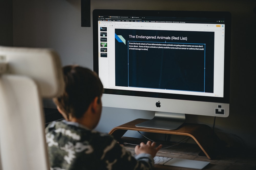 boy in white and black shirt sitting on chair in front of silver imac