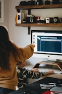 A person with long hair is pointing at a computer screen displaying information about endangered animals, while a seated child in a camouflage shirt is using the computer mouse. The room features a shelf with vintage cameras and other items above the desk.