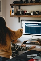 A person with long hair is pointing at a computer screen displaying information about endangered animals, while a seated child in a camouflage shirt is using the computer mouse. The room features a shelf with vintage cameras and other items above the desk.