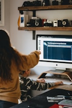 A person with long hair is pointing at a computer screen displaying information about endangered animals, while a seated child in a camouflage shirt is using the computer mouse. The room features a shelf with vintage cameras and other items above the desk.