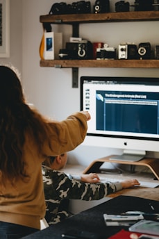 A person with long hair is pointing at a computer screen displaying information about endangered animals, while a seated child in a camouflage shirt is using the computer mouse. The room features a shelf with vintage cameras and other items above the desk.