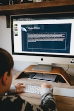A person is sitting at a desk using an iMac computer. On the screen is a presentation slide titled 'Abuse of Trees', discussing deforestation and palm oil production. The desk has a wooden stand and a keyboard. A camera and some decorative items are on a shelf above the desk.