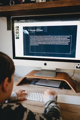 A person is sitting at a desk using an iMac computer. On the screen is a presentation slide titled 'Abuse of Trees', discussing deforestation and palm oil production. The desk has a wooden stand and a keyboard. A camera and some decorative items are on a shelf above the desk.