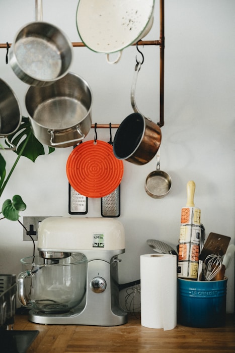 A kitchen scene featuring various utensils and appliances. Several pots and pans hang from a rack, accompanied by a red silicone trivet and kitchen graters. Below, a kitchen mixer with a glass bowl sits next to a roll of paper towels. Nearby, a container holds kitchen tools, including a rolling pin and whisk.