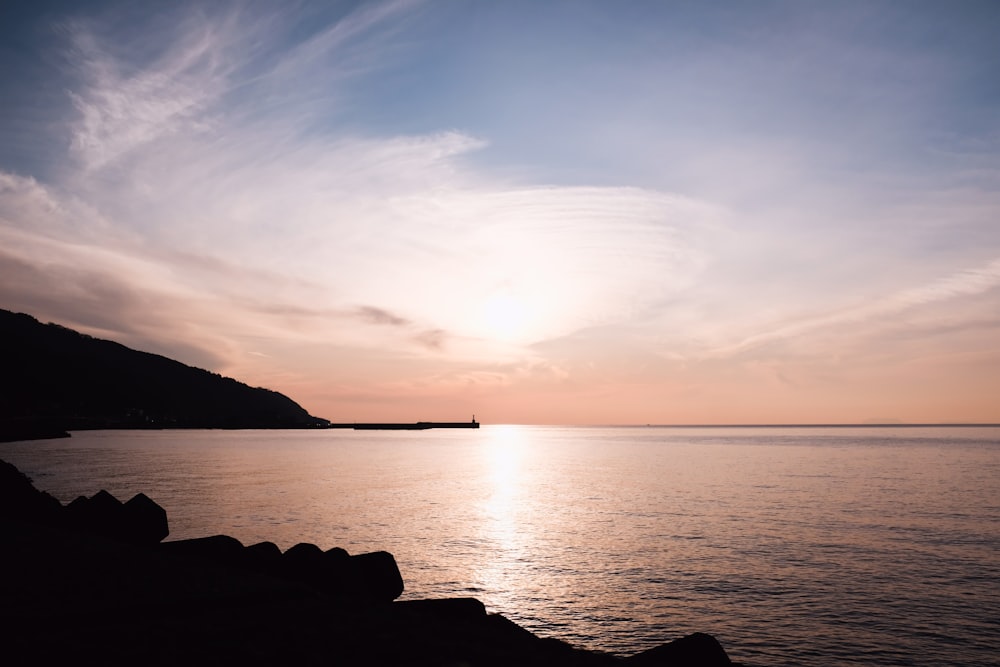 silhouette of person standing on rock near body of water during sunset