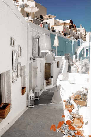 A charming outdoor street scene features narrow pathways, whitewashed buildings with blue accents, and several terraces with umbrellas. Potted plants and decorative elements add to the mediterranean aesthetic. People are seen on the balconies enjoying the setting, giving a lively yet relaxed vibe.