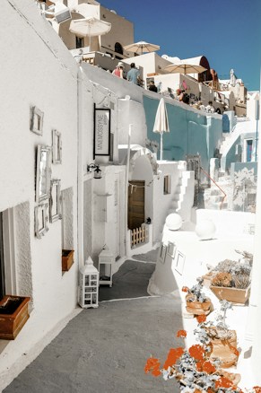 A charming outdoor street scene features narrow pathways, whitewashed buildings with blue accents, and several terraces with umbrellas. Potted plants and decorative elements add to the mediterranean aesthetic. People are seen on the balconies enjoying the setting, giving a lively yet relaxed vibe.