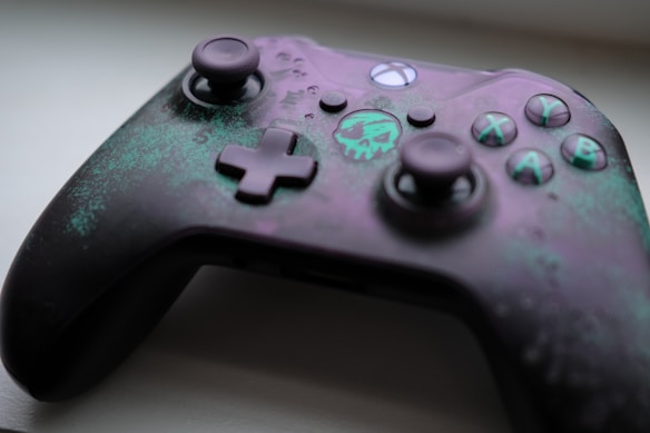 A gaming controller with a unique design featuring a skull emblem and a color scheme of purple and teal. The buttons are marked with letters and symbols, and the surface has a textured, weathered look.
