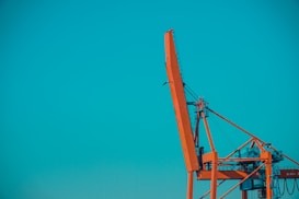 An industrial orange crane stands against a vibrant blue sky, with its complex structure of beams and cables clearly visible.