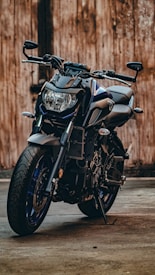 A sleek and modern motorcycle is parked on a concrete surface against a rustic wooden backdrop. The design features a robust frame with angular lines, emphasized by the bright headlamp and muscular tires. The motorcycle's body includes elements of black and blue with metallic finishes.