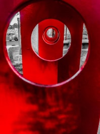 A striking image with several large red circular structures aligned in such a way that they create an optical illusion of portals receding into the distance. These structures contrast sharply with the grayscale background, where a car and some trees are visible.