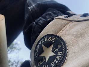 Close-up view of a beige-colored Converse Chuck Taylor All Star sneaker with the iconic circular logo. There are black laces and part of a jeans leg visible in the background.