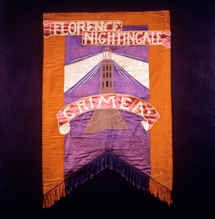 A vintage banner with a lighthouse illustration in the center, framed by orange and purple fabric. At the top, the words 'Florence Nightingale' are embroidered, and at the bottom, 'Crimea' is displayed. The banner has fringes at the bottom edge.