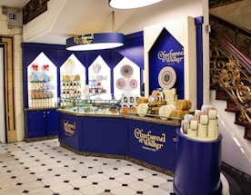 An elegantly designed store display with a royal blue and cream color scheme, showcasing a variety of round chocolate boxes and sweets. The counter is curved, with glass sections highlighting the chocolates inside, and decorative wall shelves displaying more products. A marble floor with a black and white geometric pattern complements the intricate staircase railing on the right.