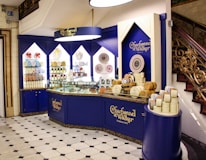 An elegantly designed store display with a royal blue and cream color scheme, showcasing a variety of round chocolate boxes and sweets. The counter is curved, with glass sections highlighting the chocolates inside, and decorative wall shelves displaying more products. A marble floor with a black and white geometric pattern complements the intricate staircase railing on the right.