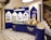 An elegantly designed store display with a royal blue and cream color scheme, showcasing a variety of round chocolate boxes and sweets. The counter is curved, with glass sections highlighting the chocolates inside, and decorative wall shelves displaying more products. A marble floor with a black and white geometric pattern complements the intricate staircase railing on the right.