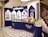 An elegantly designed store display with a royal blue and cream color scheme, showcasing a variety of round chocolate boxes and sweets. The counter is curved, with glass sections highlighting the chocolates inside, and decorative wall shelves displaying more products. A marble floor with a black and white geometric pattern complements the intricate staircase railing on the right.