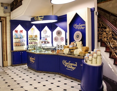 An elegantly designed store display with a royal blue and cream color scheme, showcasing a variety of round chocolate boxes and sweets. The counter is curved, with glass sections highlighting the chocolates inside, and decorative wall shelves displaying more products. A marble floor with a black and white geometric pattern complements the intricate staircase railing on the right.