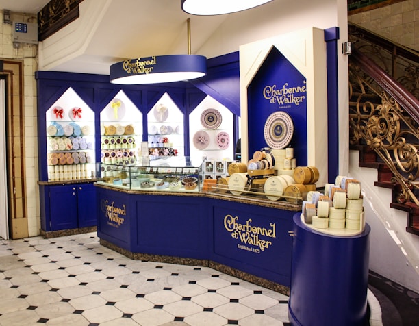 An elegantly designed store display with a royal blue and cream color scheme, showcasing a variety of round chocolate boxes and sweets. The counter is curved, with glass sections highlighting the chocolates inside, and decorative wall shelves displaying more products. A marble floor with a black and white geometric pattern complements the intricate staircase railing on the right.