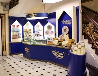 An elegantly designed store display with a royal blue and cream color scheme, showcasing a variety of round chocolate boxes and sweets. The counter is curved, with glass sections highlighting the chocolates inside, and decorative wall shelves displaying more products. A marble floor with a black and white geometric pattern complements the intricate staircase railing on the right.