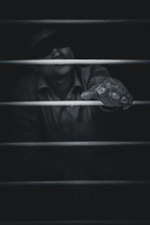 A black and white image featuring a person obscured in darkness, wearing a blindfold. The focus is on an outstretched hand reaching through bars, conveying a sense of struggle or longing. The lighting emphasizes the hand while the rest of the figure is shrouded in shadow.