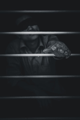 A black and white image featuring a person obscured in darkness, wearing a blindfold. The focus is on an outstretched hand reaching through bars, conveying a sense of struggle or longing. The lighting emphasizes the hand while the rest of the figure is shrouded in shadow.