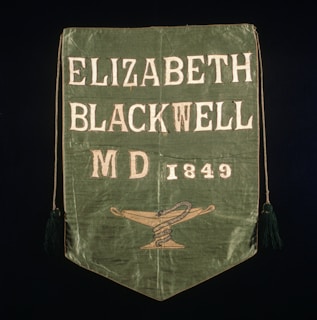 A green fabric banner with the text 'Elizabeth Blackwell MD 1849' and a symbol of a snake coiled around a staff on a lamp. The edges have tassels, and the text is embroidered in white.