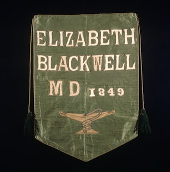 A green fabric banner with the text 'Elizabeth Blackwell MD 1849' and a symbol of a snake coiled around a staff on a lamp. The edges have tassels, and the text is embroidered in white.