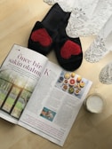 A pair of black slippers with red heart patterns is placed on a wooden floor next to a magazine and a semi-transparent lace curtain. The magazine has an article with text and images of food, possibly a healthy lifestyle or recipe section. Beside the magazine is a glass mug filled with a frothy beverage.