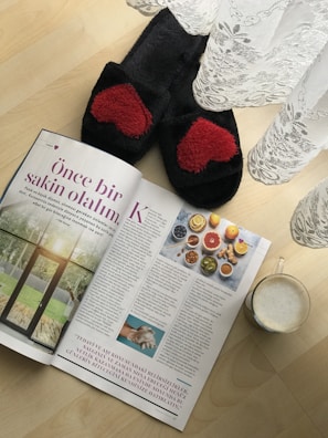 A pair of black slippers with red heart patterns is placed on a wooden floor next to a magazine and a semi-transparent lace curtain. The magazine has an article with text and images of food, possibly a healthy lifestyle or recipe section. Beside the magazine is a glass mug filled with a frothy beverage.