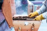 A beekeeper wearing yellow gloves is handling a wooden beehive filled with bees. Smoke is visible in the background, likely from a smoker used to calm the bees. The hive is open, revealing a large number of bees inside. The scene appears to be outdoors with a focus on the beekeeping activity.