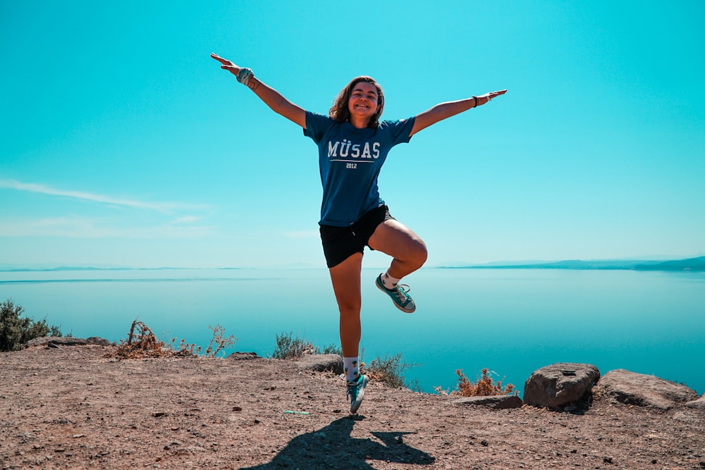 woman in blue crew neck t-shirt and black shorts jumping on brown rock near body