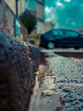 A close-up view of a wet street curb with rainwater pooled on the asphalt, focusing on the textured surface of the curb and the reflection of a blurred car in the background. The scene captures a moody, overcast ambiance with a touch of urban life.