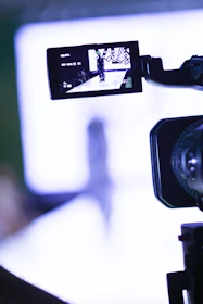 A video camera is focusing on a runway where a fashion show appears to be taking place. The camera's screen displays a person walking down the runway with bright lights and a blurred background.