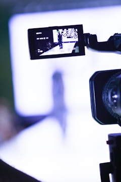 A video camera is focusing on a runway where a fashion show appears to be taking place. The camera's screen displays a person walking down the runway with bright lights and a blurred background.