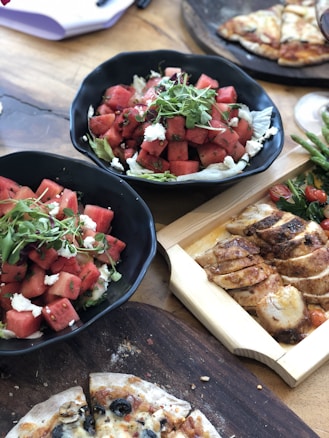 A vibrant meal featuring two black bowls filled with watermelon cubes, greens, and feta cheese on a wooden table. A rectangular wooden tray holds slices of grilled chicken. Partially visible on the side are slices of pizza topped with olives and what appears to be additional greens and tomatoes.