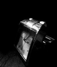 A silver wristwatch with a square face is captured in dramatic lighting. The watch has a textured dial with dark hour markers and hands, reflecting light against a dark background.