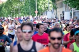 A large group of people are participating in a running event on a city street. The runners are wearing athletic attire, including shirts with numbers, sunglasses, and caps. Trees and buildings line the street in the background, and there is a mix of ages and genders among the participants.