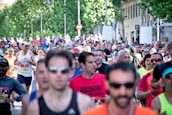 A large group of people are participating in a running event on a city street. The runners are wearing athletic attire, including shirts with numbers, sunglasses, and caps. Trees and buildings line the street in the background, and there is a mix of ages and genders among the participants.