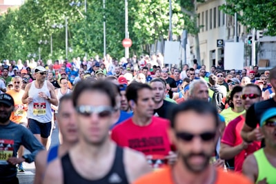 A large group of people are participating in a running event on a city street. The runners are wearing athletic attire, including shirts with numbers, sunglasses, and caps. Trees and buildings line the street in the background, and there is a mix of ages and genders among the participants.