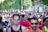 A large group of people are participating in a running event on a city street. The runners are wearing athletic attire, including shirts with numbers, sunglasses, and caps. Trees and buildings line the street in the background, and there is a mix of ages and genders among the participants.
