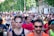 A large group of people are participating in a running event on a city street. The runners are wearing athletic attire, including shirts with numbers, sunglasses, and caps. Trees and buildings line the street in the background, and there is a mix of ages and genders among the participants.