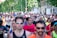 A large group of people are participating in a running event on a city street. The runners are wearing athletic attire, including shirts with numbers, sunglasses, and caps. Trees and buildings line the street in the background, and there is a mix of ages and genders among the participants.