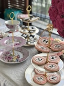 An elegant tea setting features tiered trays adorned with ornate cookies, some of which display a recognizable logo. A decorative cup and plates with floral designs add to the sophisticated aesthetic. A lush bouquet of pink roses complements the setting, adding a touch of vibrant color.