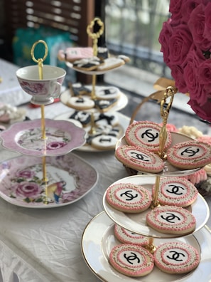 An elegant tea setting features tiered trays adorned with ornate cookies, some of which display a recognizable logo. A decorative cup and plates with floral designs add to the sophisticated aesthetic. A lush bouquet of pink roses complements the setting, adding a touch of vibrant color.