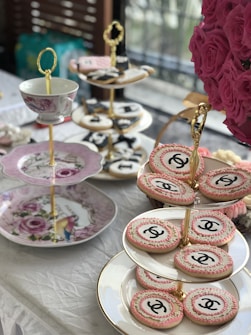 An elegant tea setting features tiered trays adorned with ornate cookies, some of which display a recognizable logo. A decorative cup and plates with floral designs add to the sophisticated aesthetic. A lush bouquet of pink roses complements the setting, adding a touch of vibrant color.