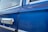 Close-up of a classic car door with a shiny chrome handle against a vibrant blue paint finish. The window features a slight reflection and is bordered by chrome trim, adding to the vintage appeal.