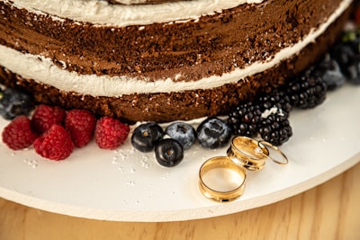 A close-up of a chocolate layer cake with cream filling, surrounded by fresh berries including raspberries, blueberries, and blackberries on a white plate. Three gold rings are placed beside the berries, adding a touch of elegance.