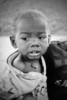 A young child with a serious expression looks to the side. The child is wearing a shirt with a monkey graphic and a necklace with an amulet. An adult’s hand gently supports the child.