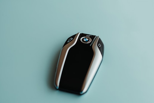 A sleek BMW car key fob with a glossy black center and metallic edges, featuring the BMW logo in the middle. The background is a smooth, light teal surface.